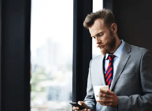 A man in a gray suit and red-striped tie stands by a window, holding a cup in one hand and looking at his phone in the other.