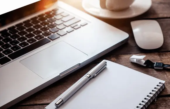 A laptop, a cup, a mouse, a notebook with a pen, and USB drives on a wooden table.