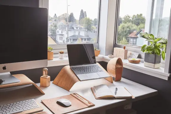 A home office setup with a computer monitor, a laptop on a stand, an open notebook with a pen, potted plants, and a window overlooking buildings and trees in the background.