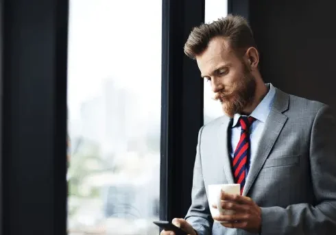 A man in a gray suit and red-striped tie stands by a window, holding a cup in one hand and looking at his phone in the other.