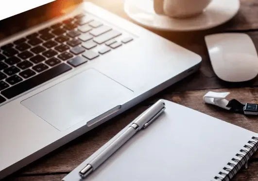 A laptop, a cup, a mouse, a notebook with a pen, and USB drives on a wooden table.