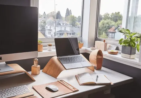 A home office setup with a computer monitor, a laptop on a stand, an open notebook with a pen, potted plants, and a window overlooking buildings and trees in the background.